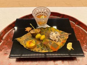 an elegant lunch presentation features a fried ginkgo leaf in fall yellow, ginkgo nuts speared with pine needles, and mushrooms, garnished artfully with dried leaves and served on a giant leaf.