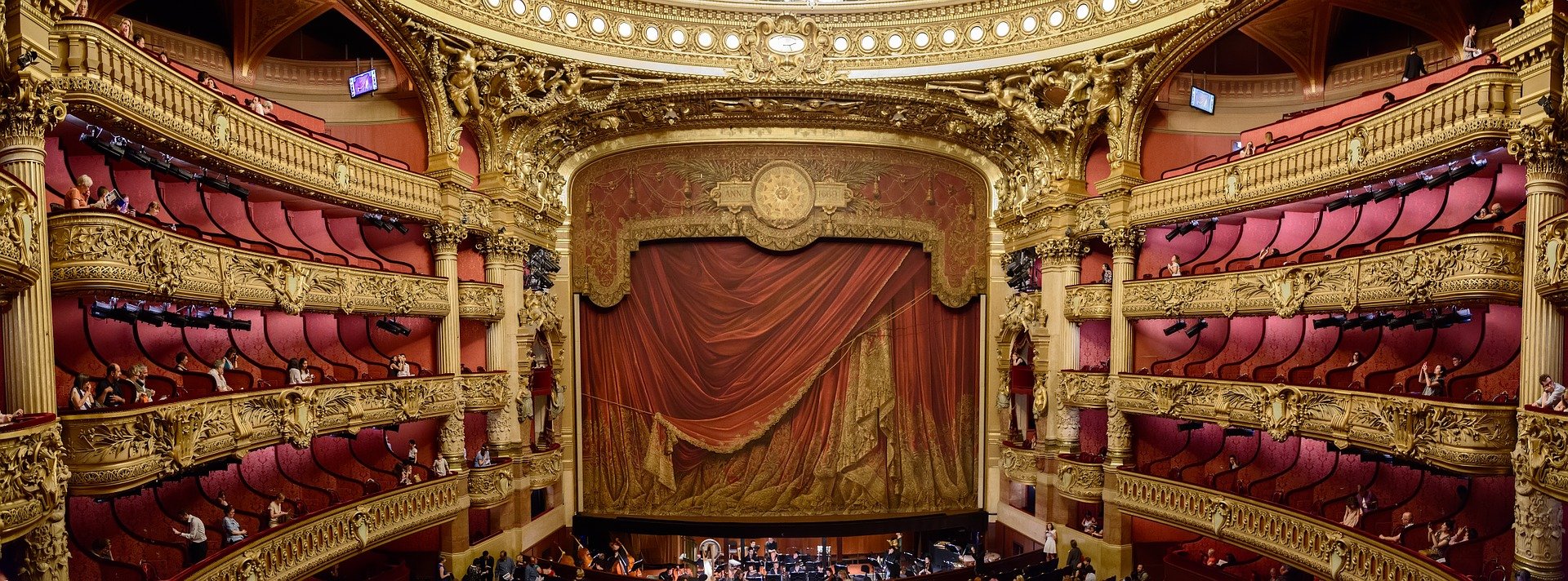 this shows a beautiful and elaborate theater with a heavy red curtain decorated with gold and 4 levels of box seats